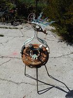 Full front view of the scrap metal sculpture showing the top-heavy circular base with gear-like structure and whimsical decorative elements including metal leaf shapes and curled wire on top.