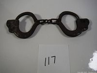 Front view of dark metal antique children's toy handcuffs with oval cuffs connected by short linked chain, displaying some rust and wear.