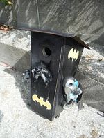 Front side view of the black wooden bird house featuring a circular entrance hole, Borussia Batman logos painted in yellow, Batman figurine mounted below the hole, and rusty tin roof with slightly bent edges.