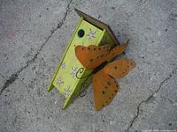 Front-side view of green bird house with decorative orange butterfly metal attached near entrance hole, showing painted floral details on sides.