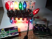Photo showing the string lights plugged in and lit with red, yellow, green, and blue bulbs in a box, with a box of replacement bulbs beside it.