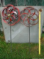 Both metal wind spinners displayed side by side against a weathered wooden backdrop with a yellow measuring stick showing height.