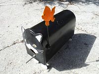 Black metal cat-shaped bird house with white painted facial features and feet, missing one whisker and tail. Orange metal fish tail on a spring rod visible.