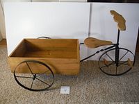 Large wooden tricycle shaped plant or display stand with a box cargo area and three metal wheels photographed on carpet with a white backing.
