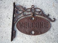 Front view showing cast iron bracket with detailed scrollwork and double-sided oval 'WELCOME' sign with rusty patina.