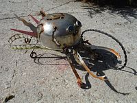 Front-right angled view of the pot-based metal sculpture with visible eyes and curled metal elements.