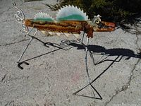 Full side view of rusty metal bug sculpture on pavement, showing saw blade wings and wire legs casting shadow