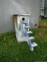 Front view of handmade birdhouse on grass with metal background, showing stairway and entrance hole.