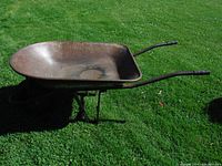 Front angled view of rust-brown metal wheelbarrow tub and black metal handles and frame, showing wear and patina on grass background.