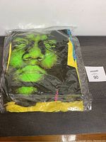 Folded yellow T-shirt showing the green graphic portrait of Notorious B.I.G. wearing a crown, sealed partially in plastic.