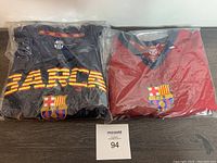Two FC Barcelona men's soccer jerseys in packaging; one black with yellow/orange 'BARCA' text, one maroon and navy, both new with tags.