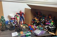 Photo showing several action figures including Spider-Man, Joker, Batman, and others, with a box of additional figures to the side.