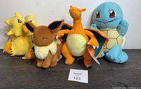 Four Pokemon plush toys: Charizard, Squirtle, Eevee, and Alakazam standing on a brown surface