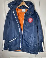 Front view of navy blue Aquatex Flotable jacket with zipper partially open showing orange quilted lining and zippered pockets.