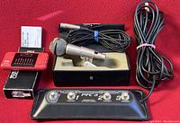 All items from the electronic music lot displayed together on a red surface: AKG microphone in black case with attached cable, red Seiko ST800 chromatic guitar tuner in original box, and black PFC3 footswitch with cable.
