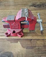Photo showing red cast iron bench vice with metal jaws and handle on wooden surface.