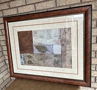 Large framed abstract art print leaning against a brick wall showing the full framed artwork with an abstract compositional design and slight damage on the frame corner.