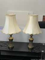 Two matching brass base table lamps with beige scalloped shades standing side by side on a black surface, cords visible.