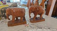 Photo showing two medium brown wooden elephant figurines sitting on rectangular wooden bases, placed on a patterned tablecloth with other items blurred in background.
