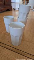 Photo shows 4 large plain white tall drinking glasses arranged on a wood floor, stacked and standing.