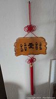 Photo showing a rectangular wooden plaque with Chinese characters referring to love, hung on a white wall with red knotted cord and tassel.