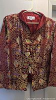 Photo of red and gold Chinese style jacket with intricate floral and swirl patterns, featuring a mandarin collar and frog button closures on a hanger.