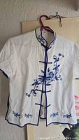White short-sleeve Chinese style top with navy blue floral embroidery, hanging on a hanger against a light background. Shows front view with mandarin collar and knotted buttons.