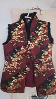 Red and gold Chinese style vest with black faux furry trim hanging on a white plastic hanger against a plain background. The vest shows detailed floral pattern with front knot closures.