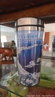 Photo shows a blue stainless steel Crater Lake National Park tumbler with lid. Positioned standing on a glass shelf with a wooden animal sculpture nearby.