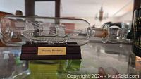 Glass ship in a bottle displayed horizontally on a separate wooden stand with brass plaque 'Priority Banking'. Bottle corked with slight discoloration inside neck from age.