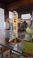 Decorative gold-plated cross adorned with clear crystals, standing on a pedestal base, shown on glass shelf with background items.