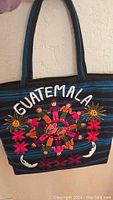 Front view showing embroidered 'GUATEMALA' text, people holding hands motif, and colorful flowers on blue and black striped fabric handbag.