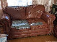 Front view of light coffee brown real leather loveseat with two seat cushions covered in silver tape for damage protection. Shows armrests with nailhead trim and worn cushions.