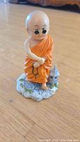 Front view of a small ceramic monk figurine wearing orange robe and round sunglasses, holding prayer beads, standing on rocky detailed base.