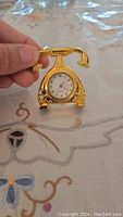 Photo of small gold-coloured clock shaped like a vintage telephone being held by fingers to show size, placed over a decorative embroidered fabric.
