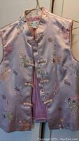 Photo of a pink traditional Chinese vest hanging, showing front details including floral embroidery and frog button closures.