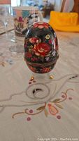 Closed view of black egg-shaped box with red and pink rose painted decoration on fabric tablecloth