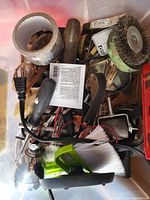 Plastic bin containing assorted miscellaneous tools such as wire brushes, adhesive tape, handheld brushes, electrical plug and cord, and metal scraping tools.