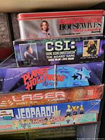 Photo of stack of seven board games including Desperate Housewives Dirty Laundry Game in metal tin, CSI The Board Game, Planet Hollywood, Laser, Jeopardy Monopoly, and two others.