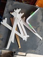 Photo of seven white and one beige metal brackets scattered on a black flat surface showing their shape and condition.