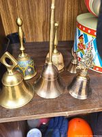 Six brass bells on wood shelf in various shapes and sizes with decorative elements