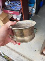 Photo of a small brass bowl being held in hand, showing tarnished surface and two small handles on opposite sides.