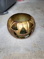 Brass hollow pumpkin with carved openings forming a jack-o'-lantern face, photographed on concrete floor.
