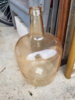 Large glass jug with a long neck and slightly amber tint, photographed outdoors on a concrete surface near a white step and metal pole.