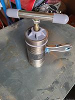 Handheld manual grinder placed on a surface, showing the crank handle, clamp lever, and metal body details.