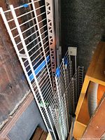 Photo showing multiple white wire grid shelving panels stacked vertically alongside metal vertical support beams with slotted holes for shelf mounting. Blue and green plastic clips are attached