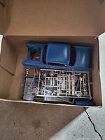 Top-down photo of open cardboard box containing a blue painted model car body with red interior visible, along with several grey plastic parts sprues for assembly.