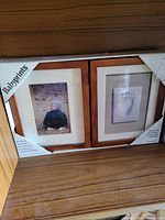 Original boxed set containing two wooden baby-themed picture frames with white mats; one frame displaying a baby photo and the other a baby footprint illustration.