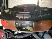 Front and top view of black JVC RC-ST1 radio and cassette player showing control buttons, speakers, digital display, and large tuning and volume knobs.