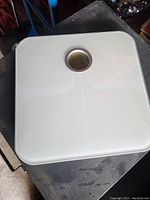 White square bathroom scale with a circular dial in the center, photographed on a dark surface.